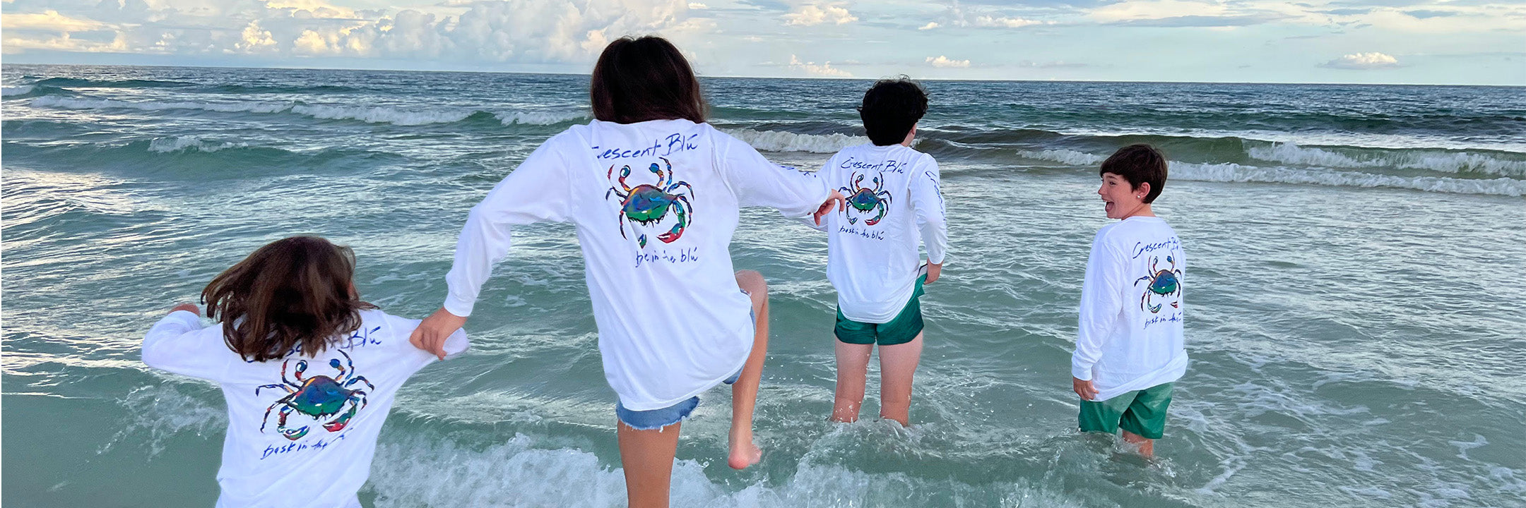 Four kids wearing long sleeve Crescent Blu Signature Crab long sleeve white t-shirt jump in the ocean waves.