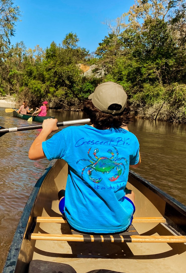 A boy wearing a brown hat and a Crescent Blú t-shirt paddles his canoe in the Bogue Falaya River in Covington, Louisiana.