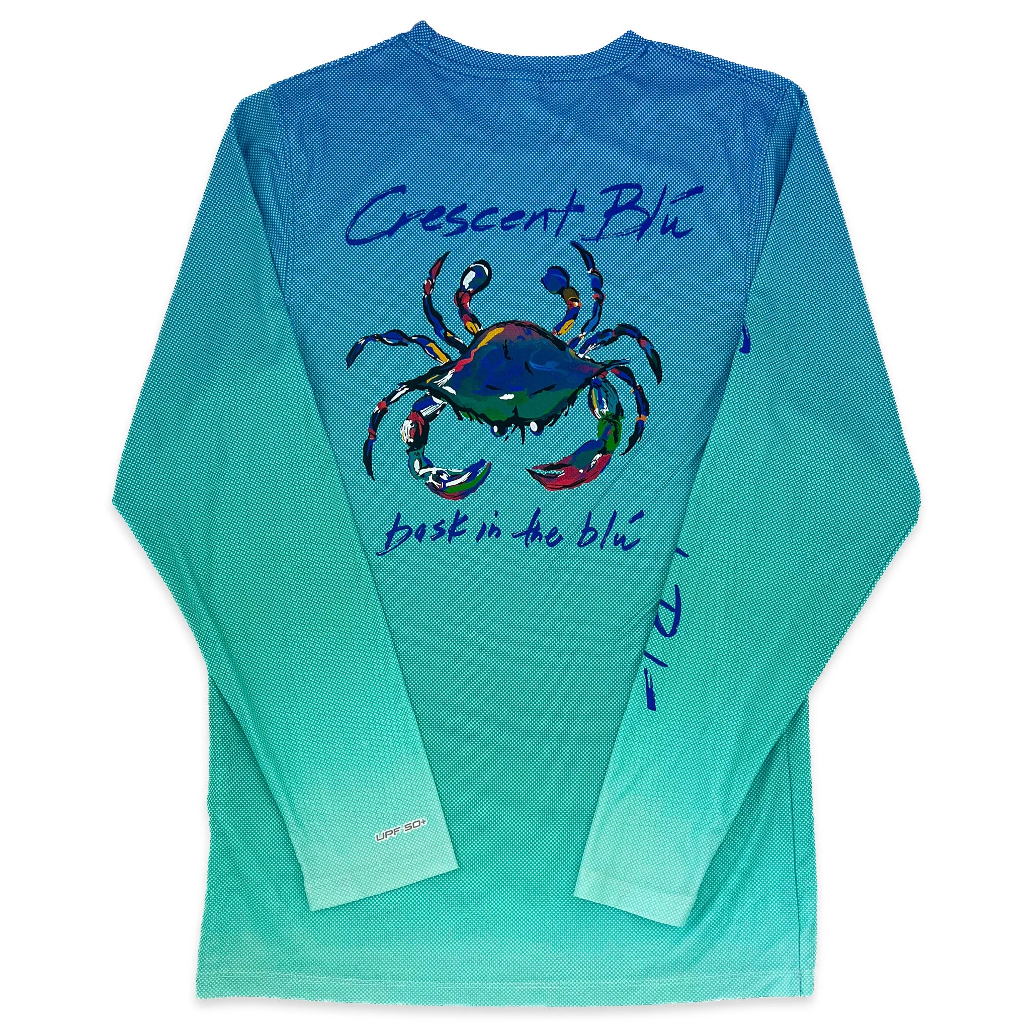 A blue crab is on the back of a long sleeve shirt.  The background of the shirt is  gradient color from  blue at the top to teal at the bottom. "Crescent Blu" is written above the crab and "bask in the blu" is written below it.