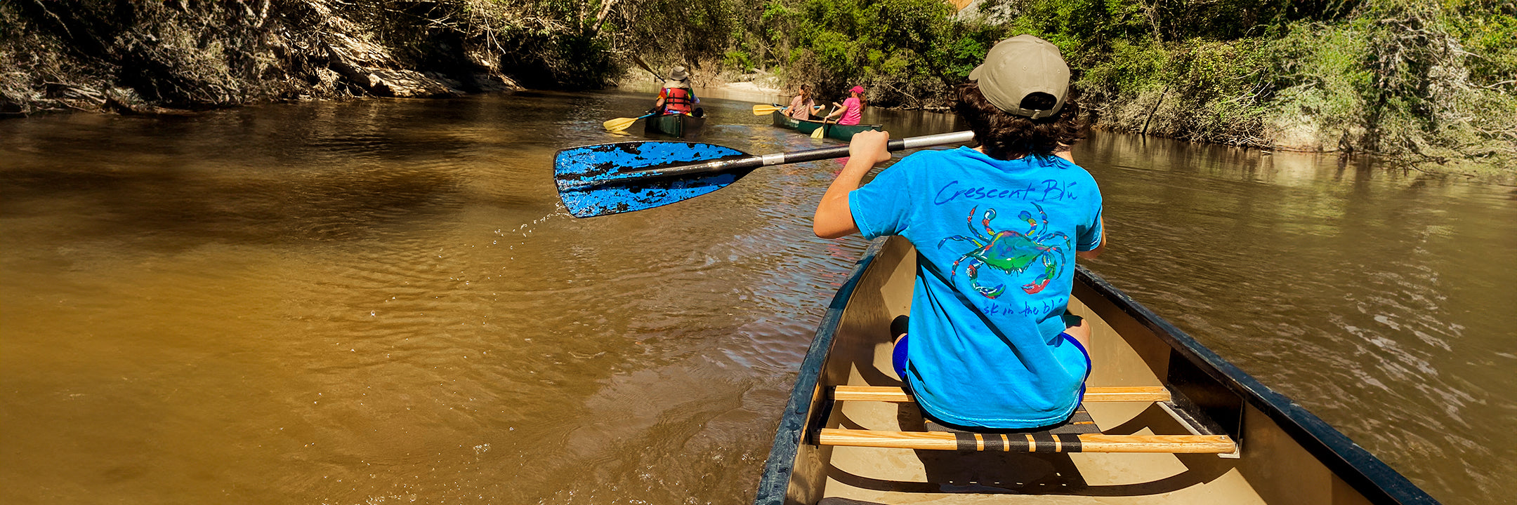A boy wearing a brown hat and a Crescent Blú t-shirt paddles his canoe in the Bogue Falaya River in Covington, Louisiana.