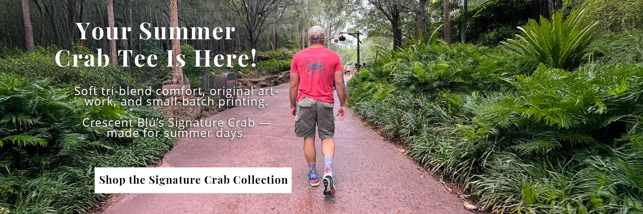 "Man walking in a Signature Crab red tri-blend t-shirt by Crescent Blú, surrounded by lush summer greenery. Back graphic shows original blue crab artwork and 'bask in the blú' tagline."