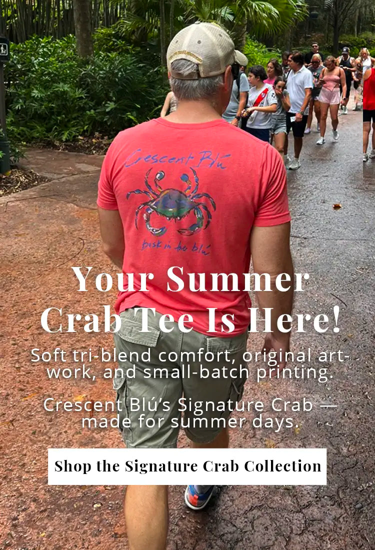 Man wearing a red Crescent Blú Signature Crab tri-blend t-shirt walking through a tree-lined path. Shirt features original crab artwork and 'bask in the blú' message on back.