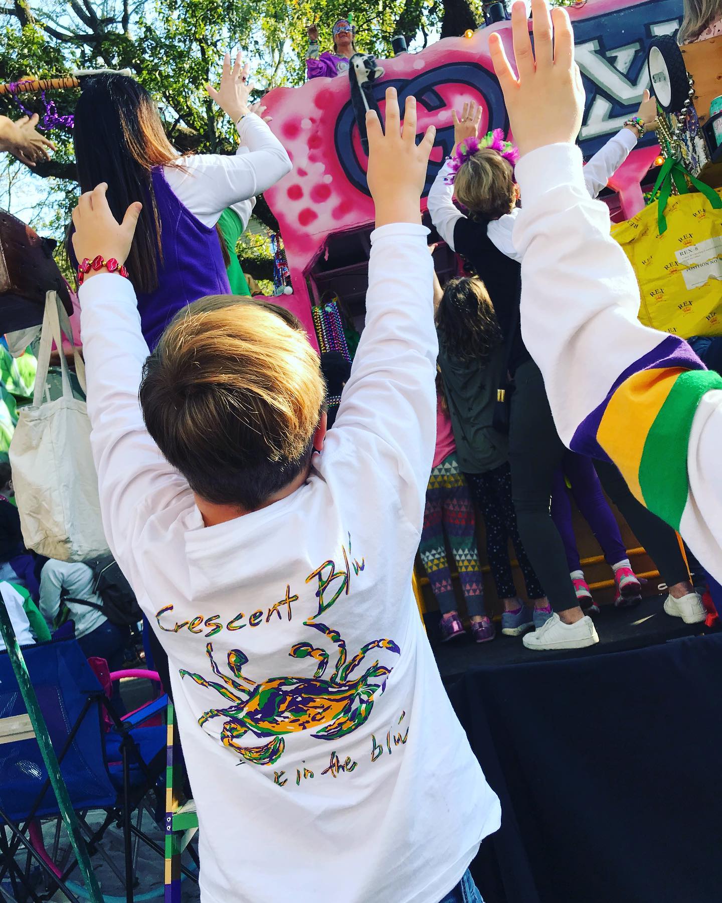 Young boy with arms raised at a Mardi Gras parade yelling for beads and wearing a long sleeve white tee showcasing a Mardi Gras colored Crescent Blu crab logo and "Bask in the Blu" written" underneath on the shirt