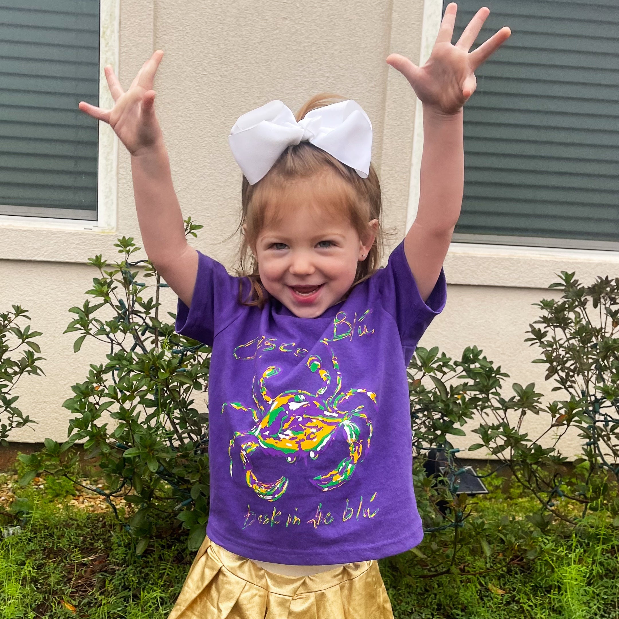 young girl with a white bow in her hair, stand infant of a house with her arms outstretched wearing a crescent blu Mardi Gras t-shirt and a gold skirt.