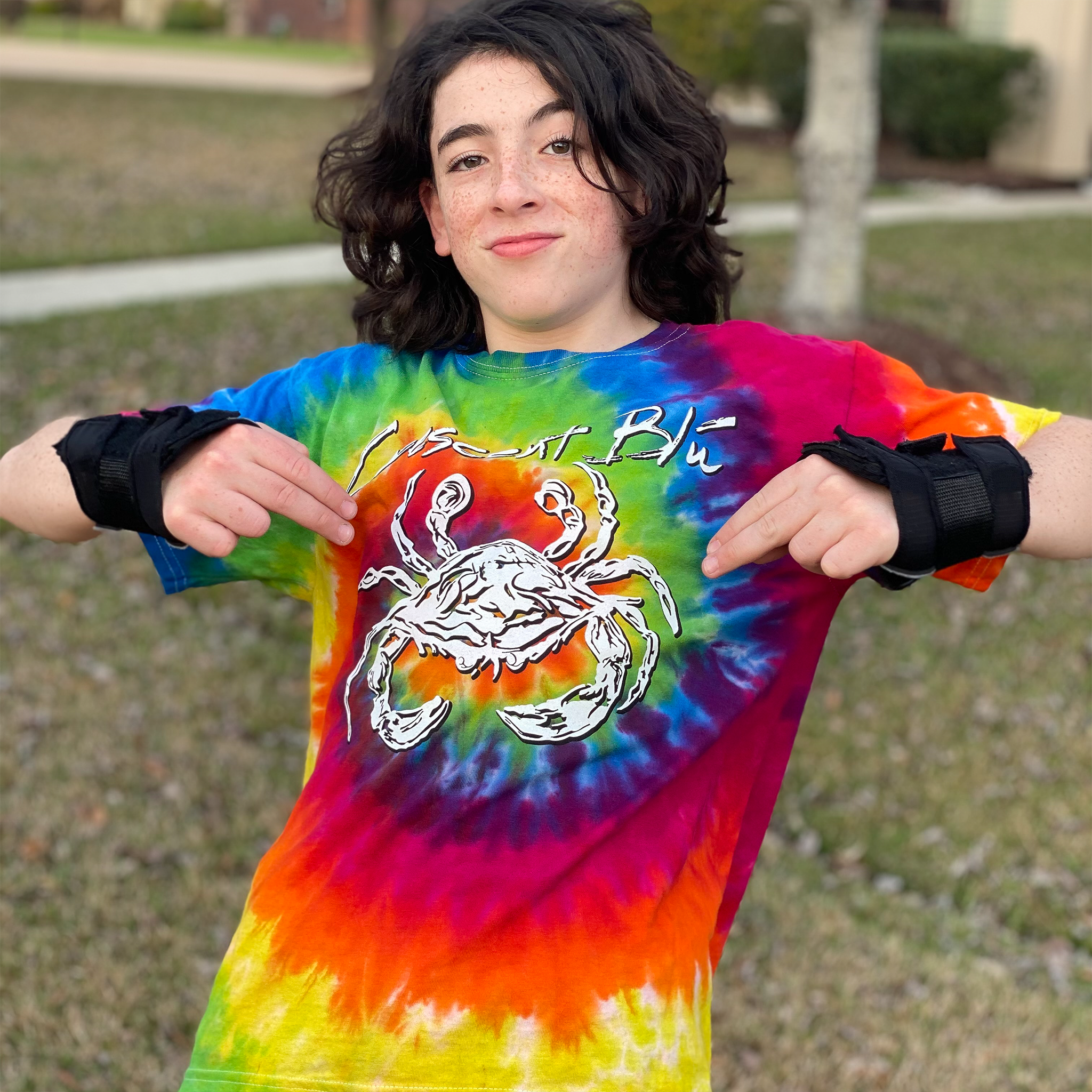 A boy wearing wrist gaurds point to a white crab in the front of his tie dyed tee shirt. 