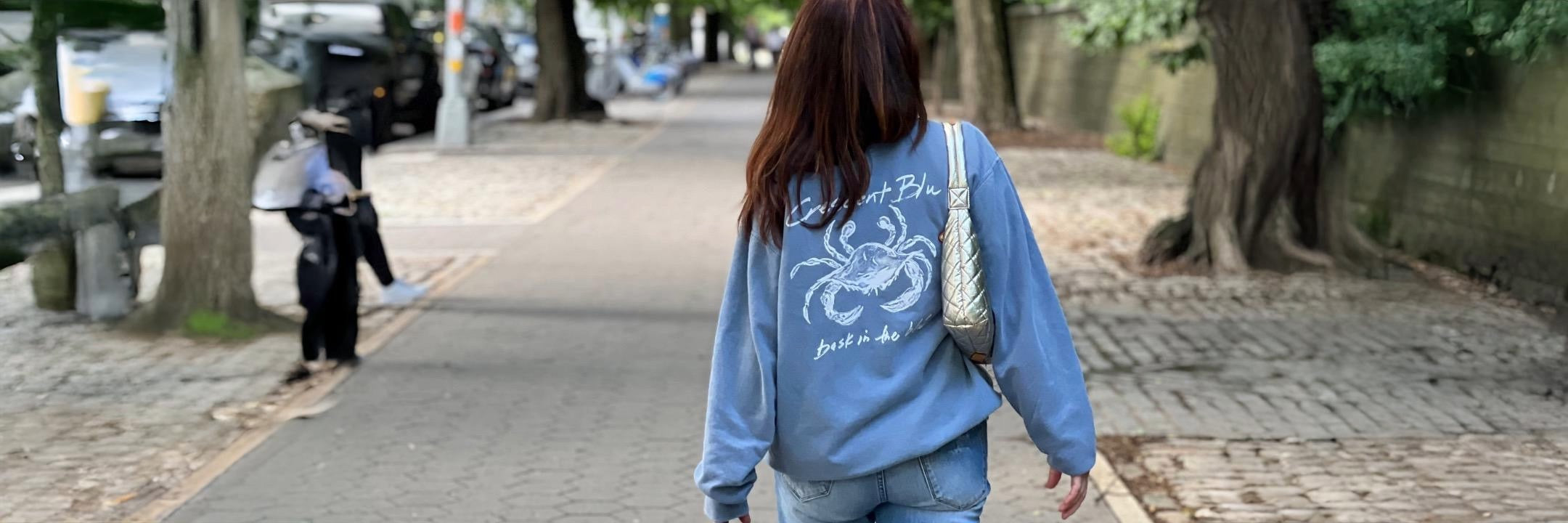woman in a Crescent Blu sneaux crab blue sweatshirt walks on the street of NYC with a gold purse 