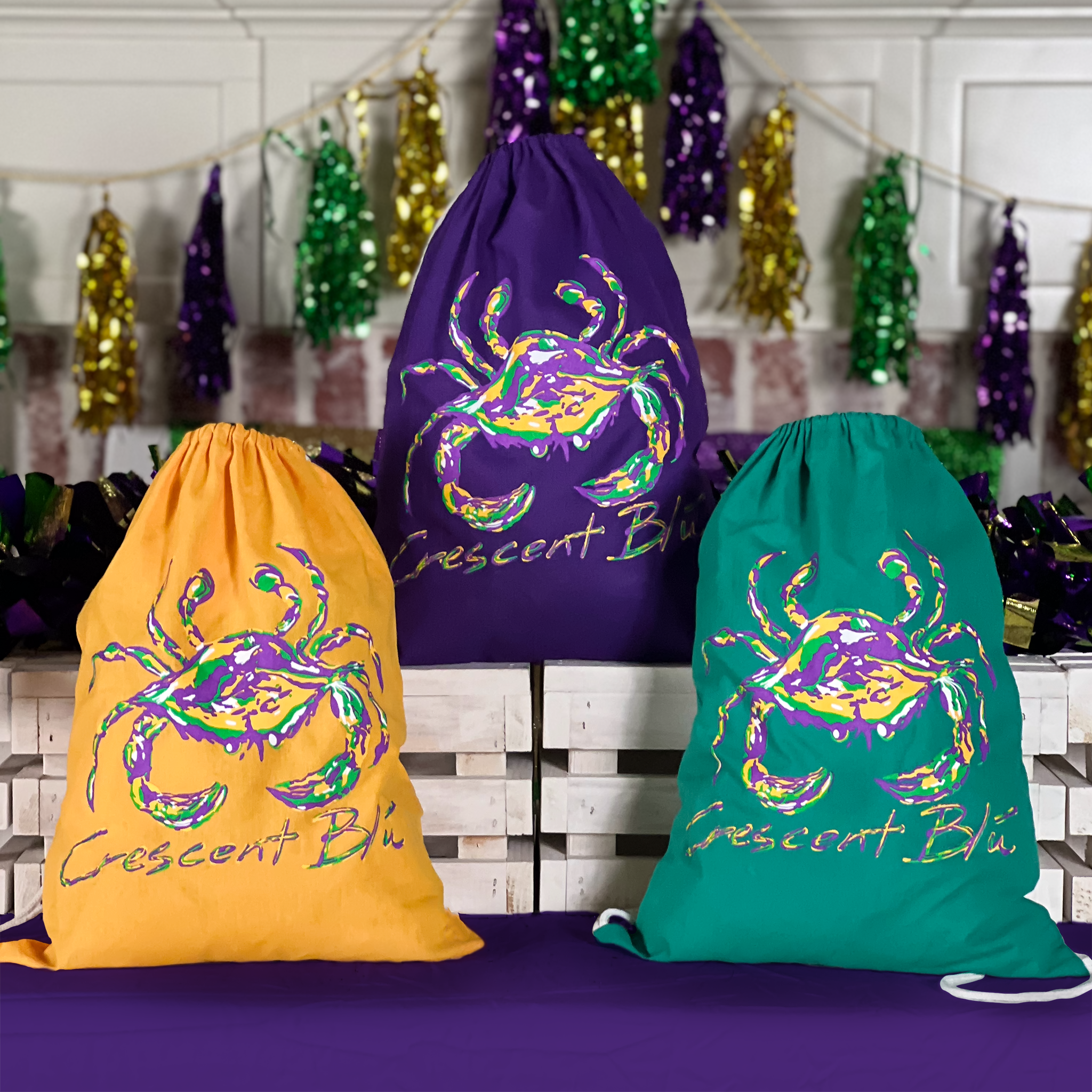 Three Mardi Gras Crab cinch sack-style  backpacks, a gold and green on a purple table and a purple backpack on white crates with  Mardi Gras tinsel hung above a fireplace.