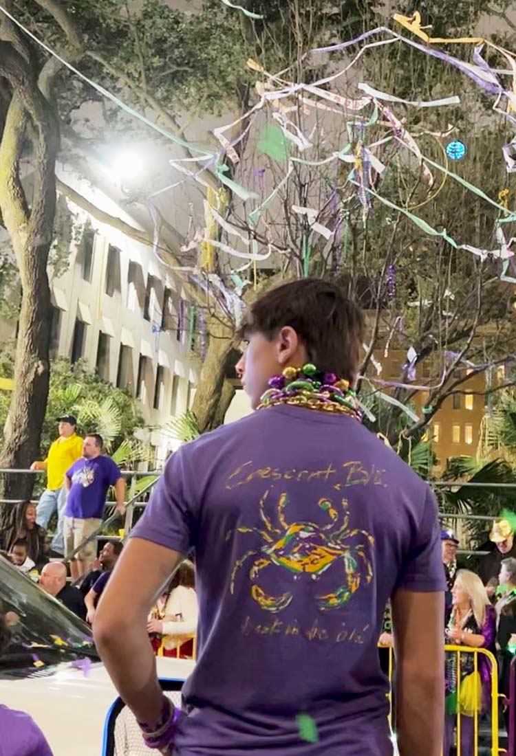 A guy wearing a Mardi Gras crab shirt stands beneath oaks trees streaming with colorful toilet paper. 