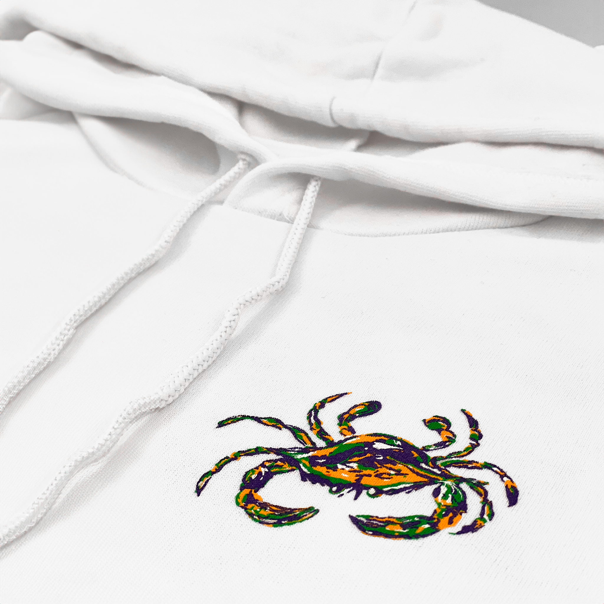 A close up of the front left chest of a white Mardi Gras Crab logo Sweatshirt with a hood and white drawcord.