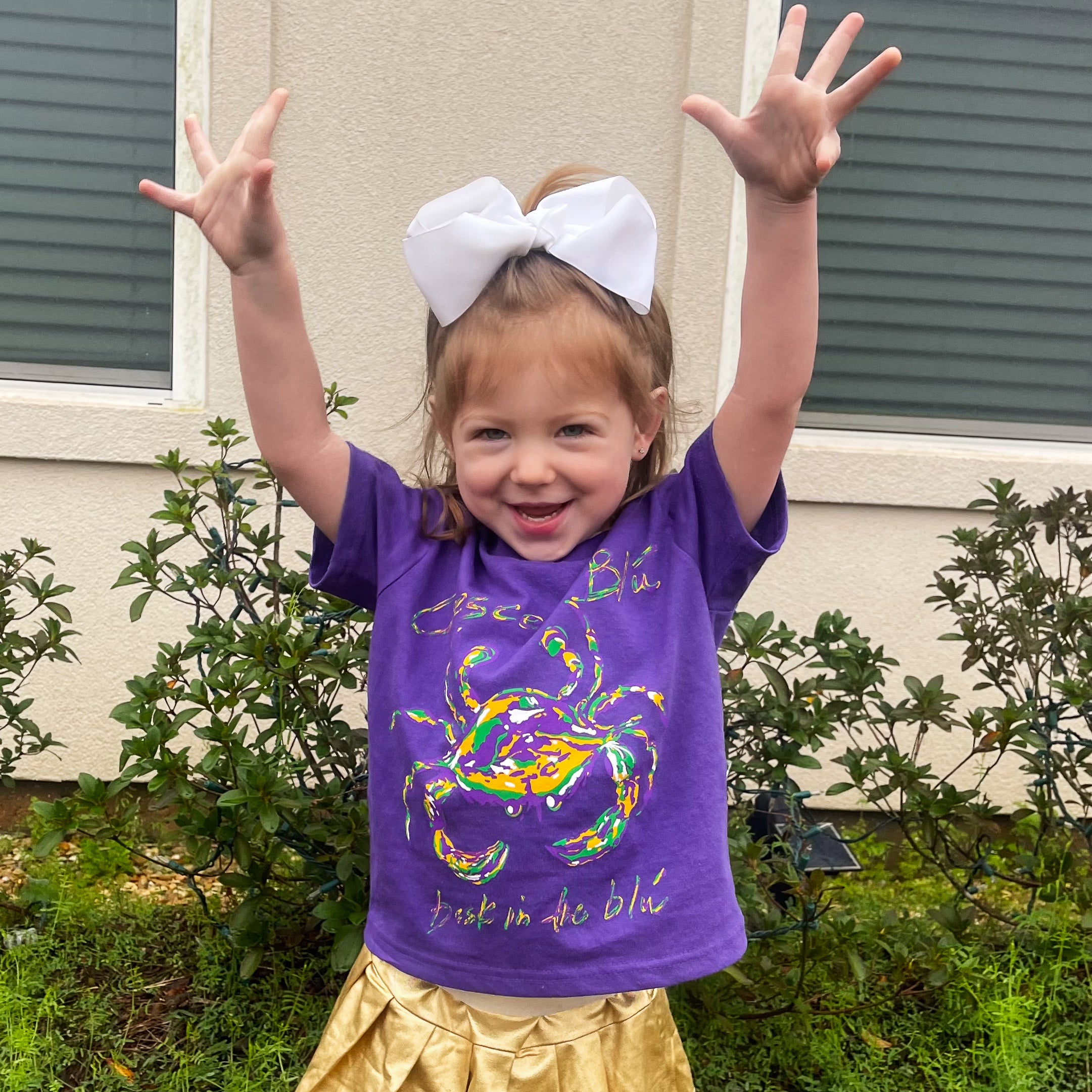 A little girl in a gold skirt and a purple Mardi Gras shirt stand in front of a house with her hands up in the air.