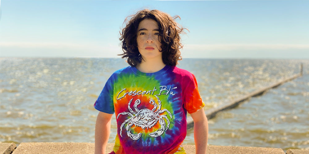 A teen boy with long hair in a tie dye shirt with a Crescent Blu logo crab on its front sits with a view of a lake behind him