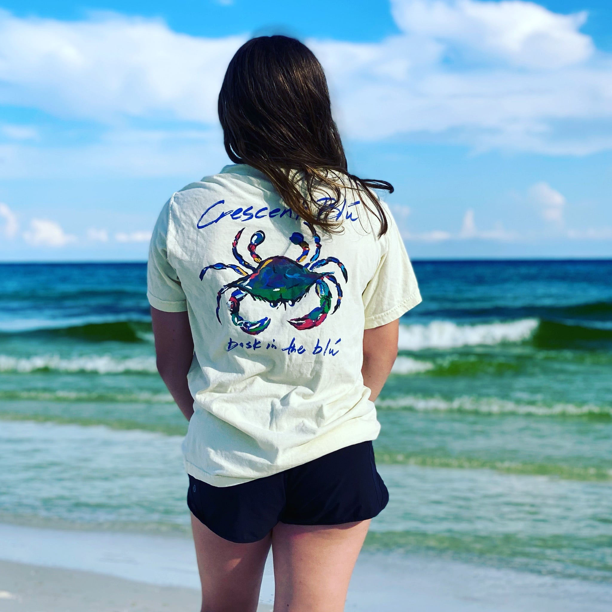 A woman in the short sleeve t-shirt with a blue crab on the back stands facing the blue waters of the ocean.