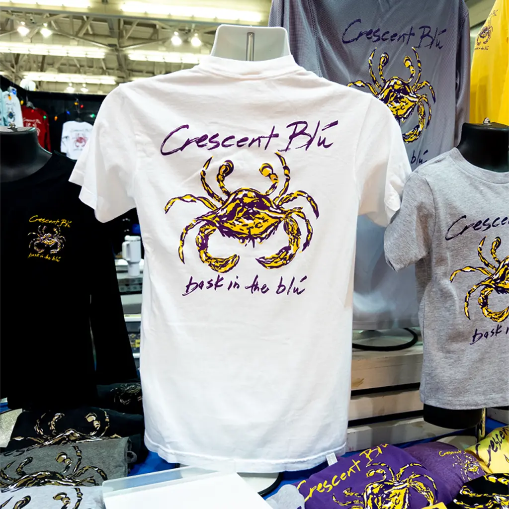White t-shirt with crab design and 'Crescent Blu' text on a mannequin, surrounded by other Crescent Blú shirts.