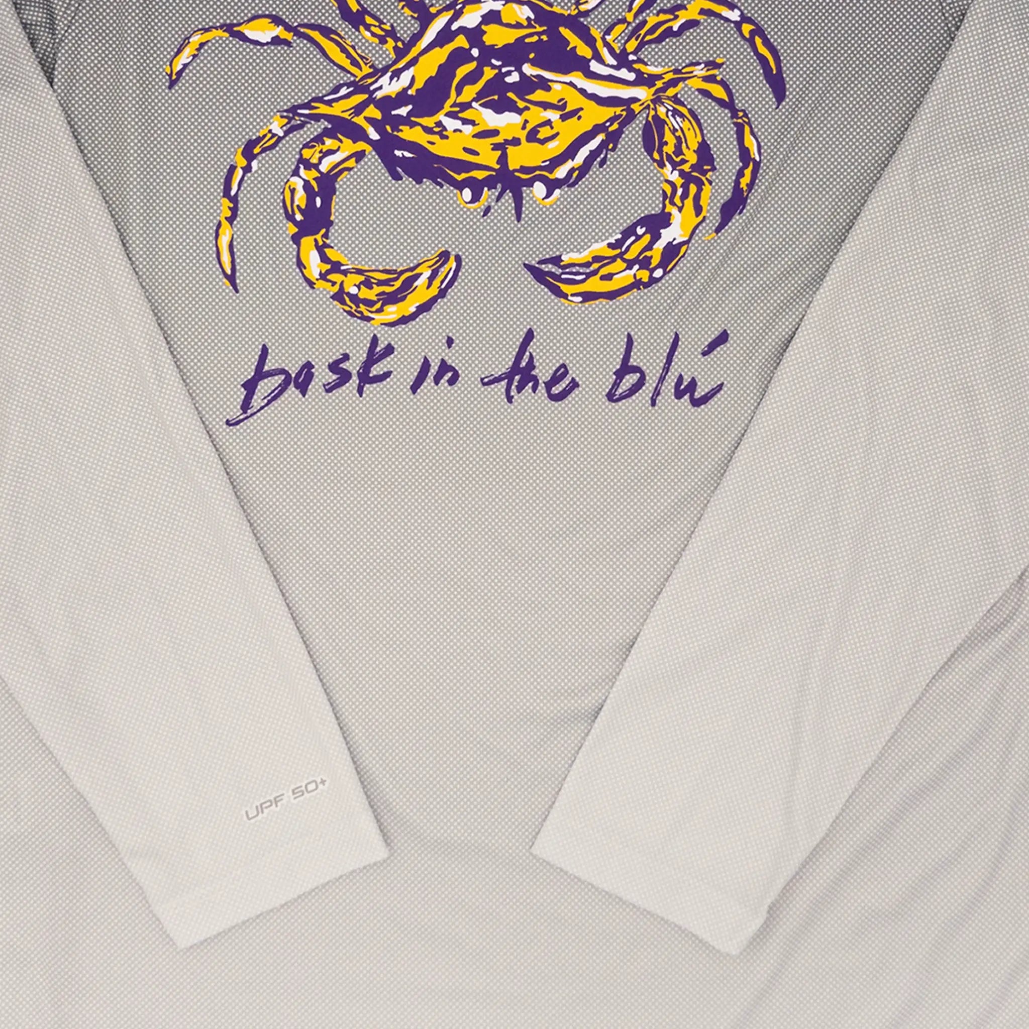 Close-up of a long-sleeve performance shirt laid flat, showing a large yellow and purple crab graphic above the words 'bask in the blu' in purple and UPF+ 50 written in grey on the near the right sleeve hem