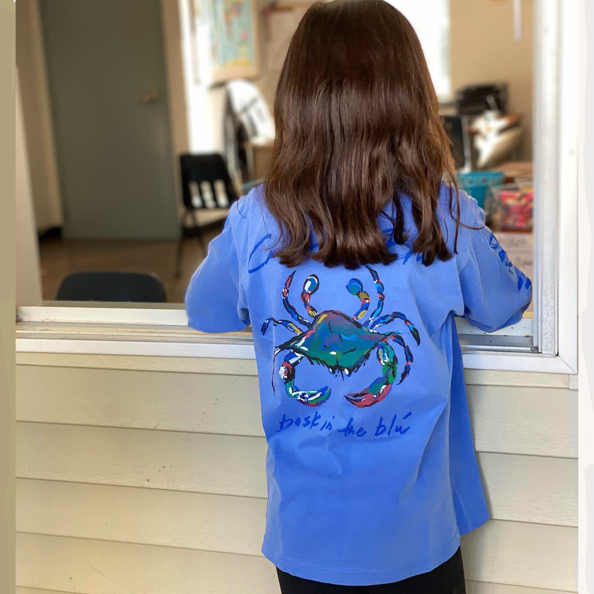 A girl in a long sleeve flo blue Crescent Blu t-shirt stand at a window. 