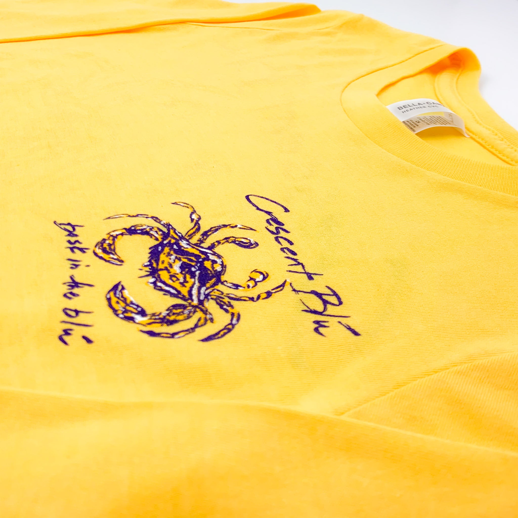 Heather yellow gold tee shirt. Crescent Blu is written in purple on the left chest above a purple, gold, and white blue crab with the tag line bask in the pule written in purple below the crab.